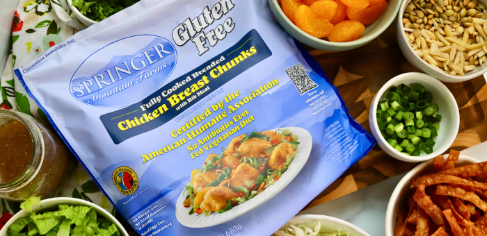 Springer Mountain Farms fully cooked gluten-free chicken breast chunks in packaging surrounded by fresh salad ingredients including lettuce, green onions, almonds, mandarin oranges, and crispy wonton strips.