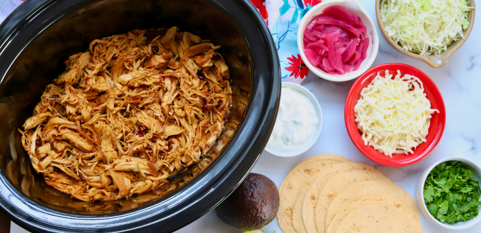 Shredded honey chipotle chicken in a slow cooker surrounded by taco toppings like cheese, cabbage, and avocado — flavorful crockpot chicken taco meal prep with Springer Mountain Farms chicken.