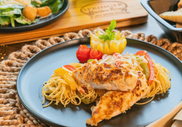 Creamy Chicken Scampi served over angel hair pasta with sautéed bell peppers, garnished with carved vegetables on a black dinner plate, paired with salad and toasted bread in the background on a rustic table.