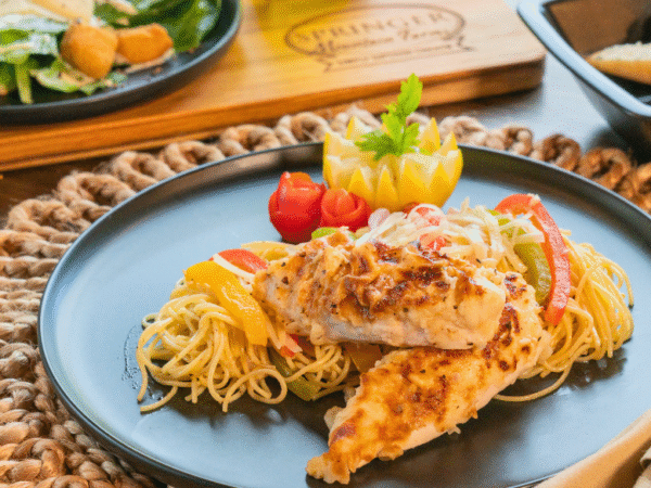 Creamy Chicken Scampi served over angel hair pasta with sautéed bell peppers, garnished with carved vegetables on a black dinner plate, paired with salad and toasted bread in the background on a rustic table.