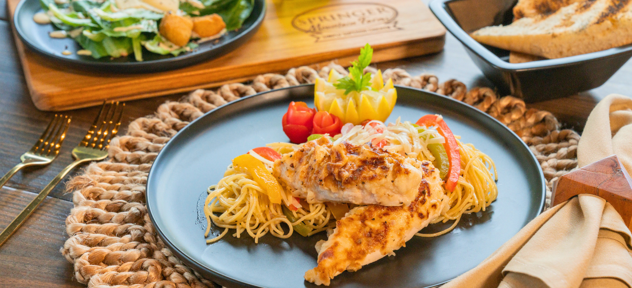 Creamy Chicken Scampi served over angel hair pasta with sautéed bell peppers, garnished with carved vegetables on a black dinner plate, paired with salad and toasted bread in the background on a rustic table.