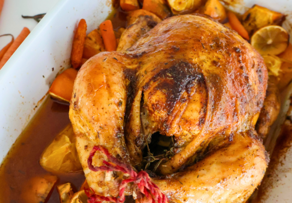 Golden brown citrus herb roasted whole Springer Mountain Farms chicken resting on carrots, sweet potatoes, lemons, and fresh herbs in a white baking dish, showcasing crispy skin and juicy pan drippings.