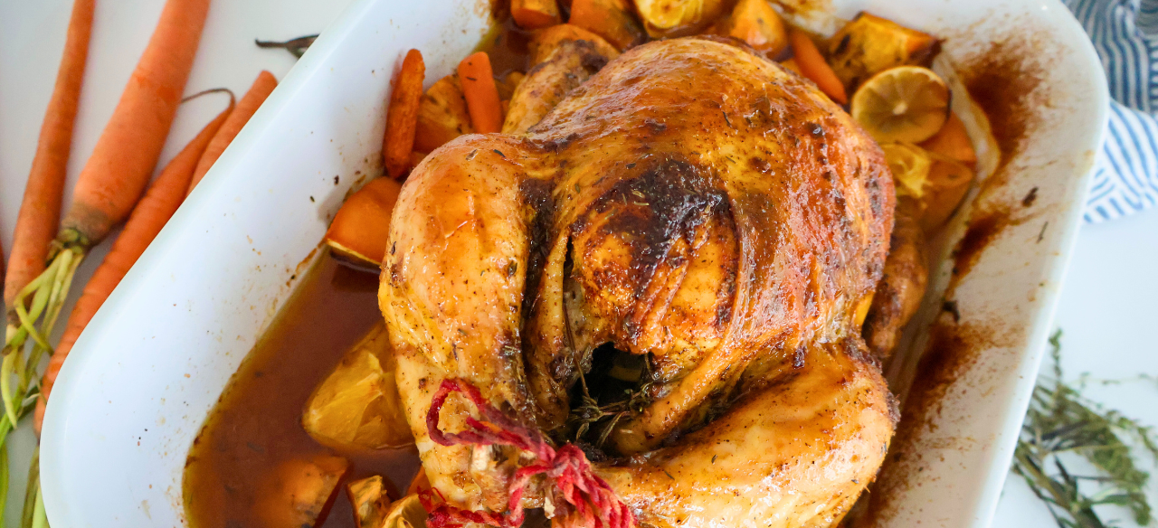Golden brown citrus herb roasted whole Springer Mountain Farms chicken resting on carrots, sweet potatoes, lemons, and fresh herbs in a white baking dish, showcasing crispy skin and juicy pan drippings.