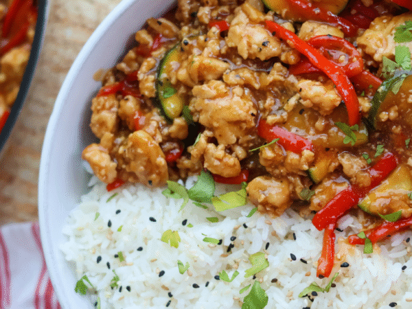 Savory ground chicken stir fry with red bell peppers, zucchini, and a flavorful sauce served over fluffy white rice with sesame seeds and fresh herbs.