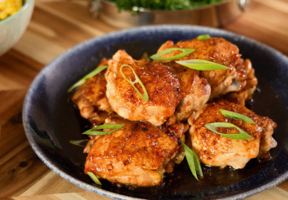 Juicy honey garlic glazed chicken thighs topped with sliced green onions on a dark plate, served with roasted potatoes and corn for a cozy homemade dinner.