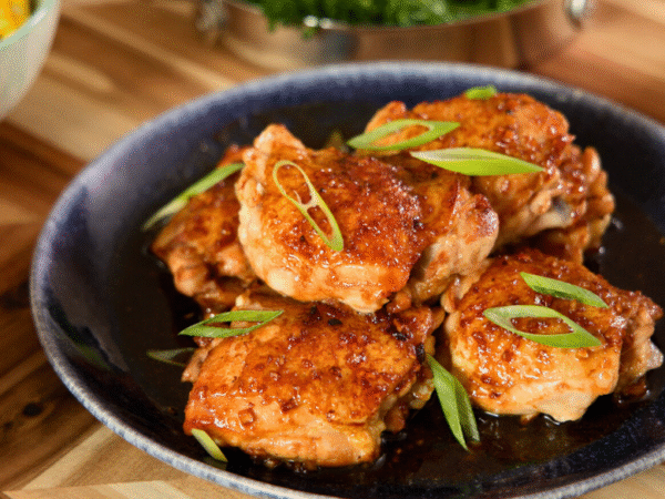 Juicy honey garlic glazed chicken thighs topped with sliced green onions on a dark plate, served with roasted potatoes and corn for a cozy homemade dinner.