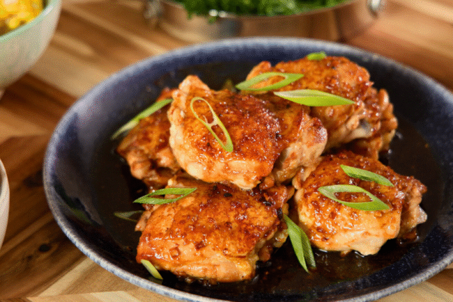 Juicy honey garlic glazed chicken thighs topped with sliced green onions on a dark plate, served with roasted potatoes and corn for a cozy homemade dinner.