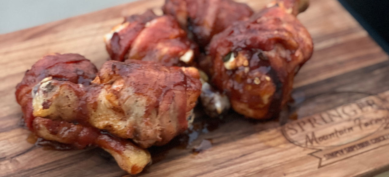 Grilled jalapeño popper stuffed chicken drumsticks wrapped in crispy bacon, glazed with barbecue sauce, served on a Springer Mountain Farms wooden board.