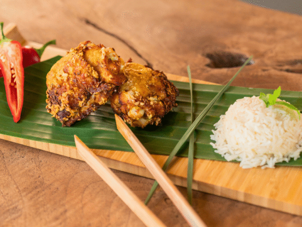 Malaysian Fried Chicken Thighs (Ayam Goreng) served on a banana leaf-lined wooden tray with crispy golden chicken pieces, sliced red chili, a mound of steamed white rice, and chopsticks, styled on a rustic wooden table.