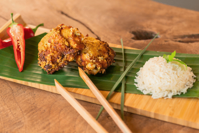 Malaysian Fried Chicken Thighs (Ayam Goreng) served on a banana leaf-lined wooden tray with crispy golden chicken pieces, sliced red chili, a mound of steamed white rice, and chopsticks, styled on a rustic wooden table.