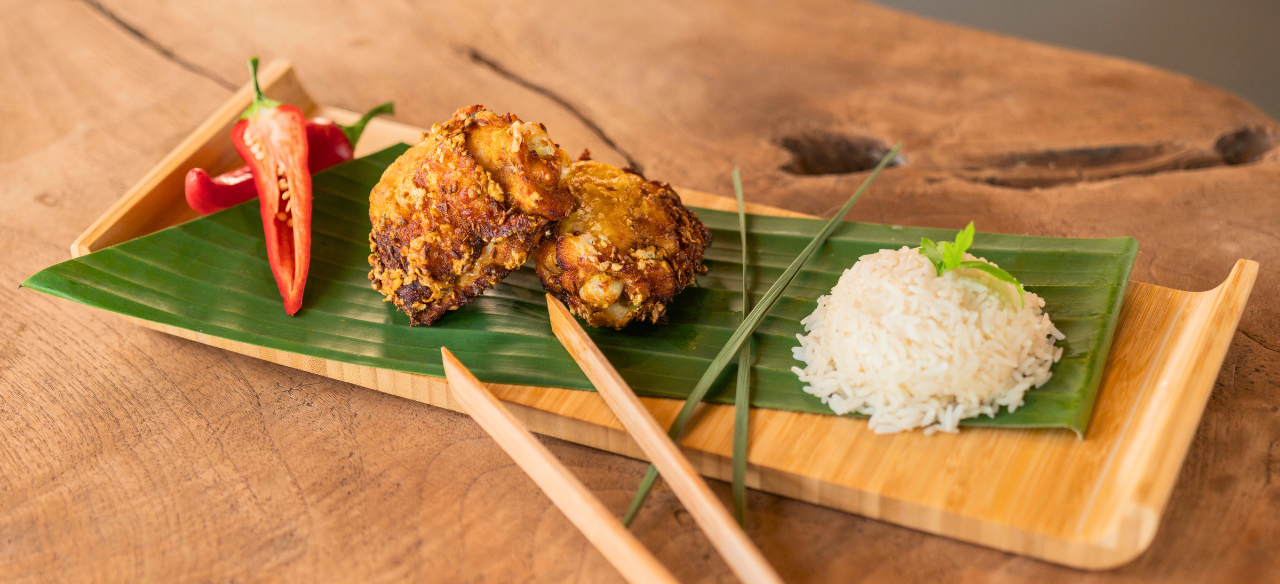 Malaysian Fried Chicken Thighs (Ayam Goreng) served on a banana leaf-lined wooden tray with crispy golden chicken pieces, sliced red chili, a mound of steamed white rice, and chopsticks, styled on a rustic wooden table.