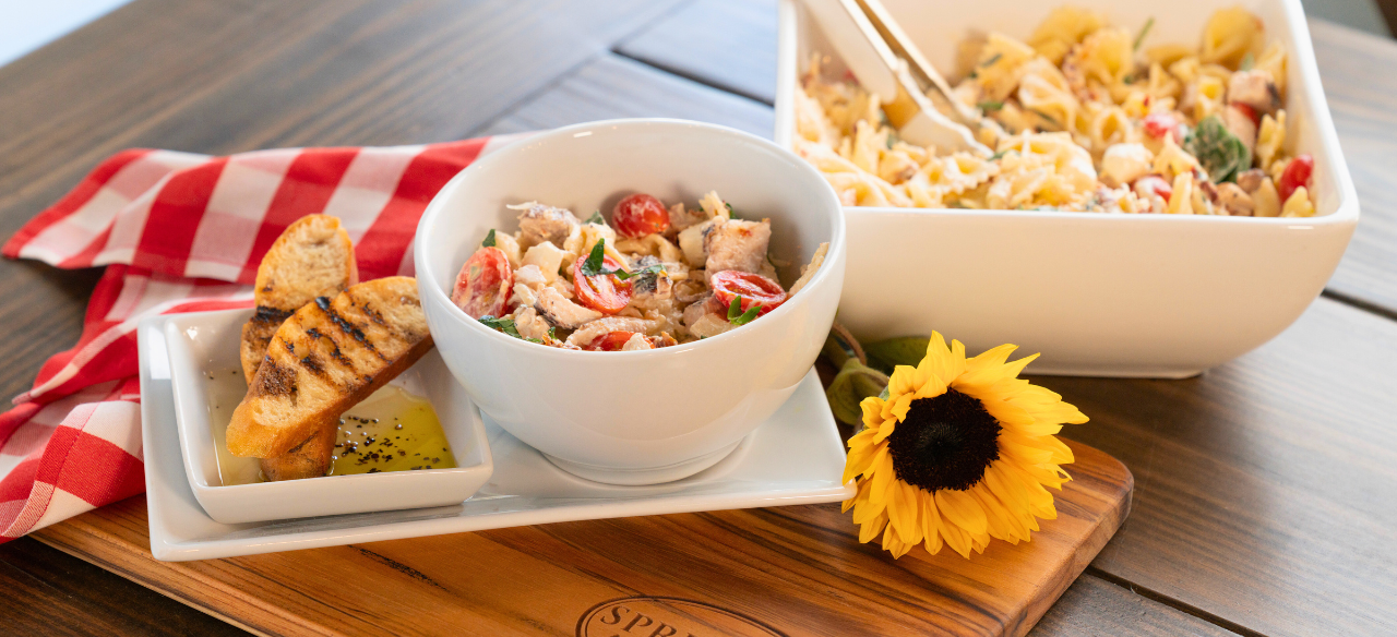 Marry Me Chicken Pasta Salad served in a white bowl with cherry tomatoes, fresh herbs, and tender chicken, accompanied by grilled baguette slices and olive oil dip on a Springer Mountain Farms wooden cutting board, set on a rustic table with a red gingham napkin and a sunflower for décor.