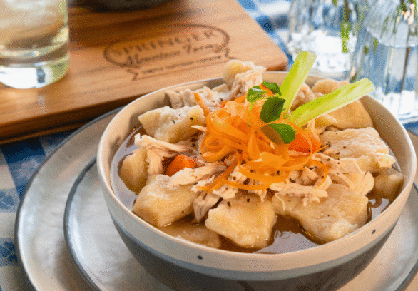 Bowl of old-fashioned Southern chicken and dumplings made with tender Springer Mountain Farms shredded chicken, fluffy dumplings, carrots, and rich broth, garnished with carrot ribbons and celery on a checkered tablecloth.