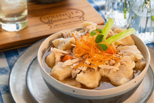 Bowl of old-fashioned Southern chicken and dumplings made with tender Springer Mountain Farms shredded chicken, fluffy dumplings, carrots, and rich broth, garnished with carrot ribbons and celery on a checkered tablecloth.