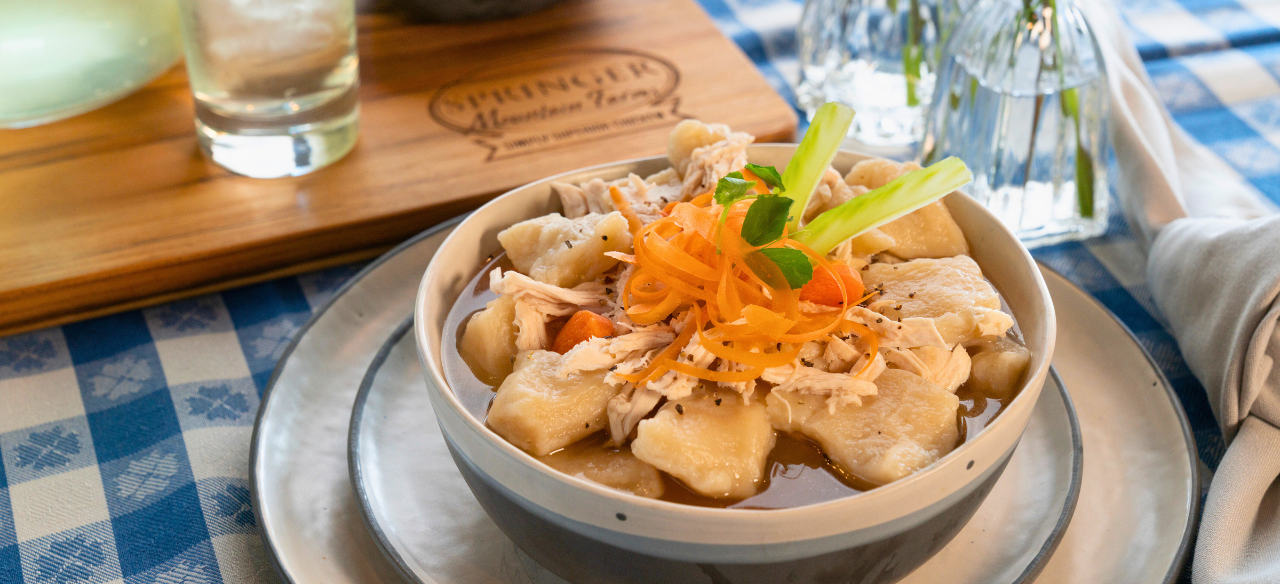 Bowl of old-fashioned Southern chicken and dumplings made with tender Springer Mountain Farms shredded chicken, fluffy dumplings, carrots, and rich broth, garnished with carrot ribbons and celery on a checkered tablecloth.