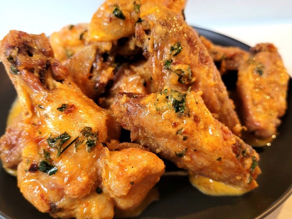 A close-up shot of crispy baked chicken wings coated in rich, golden cowboy butter, served on a black plate. The wings have a crunchy, golden-brown exterior, generously covered in a buttery glaze infused with garlic, herbs, and spices. Finely chopped parsley and crushed red pepper flakes are scattered on top, adding pops of color. Some of the warm, melted butter pools on the plate, emphasizing the dish’s rich and savory appeal. The background is minimalistic, drawing full attention to the wings' glistening texture and appetizing presentation.