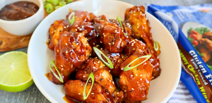 Fully cooked gluten-free savory chicken wings from Springer Mountain Farms, air fried and tossed in teriyaki sauce, served in a white bowl with sliced green onions and sesame seeds.