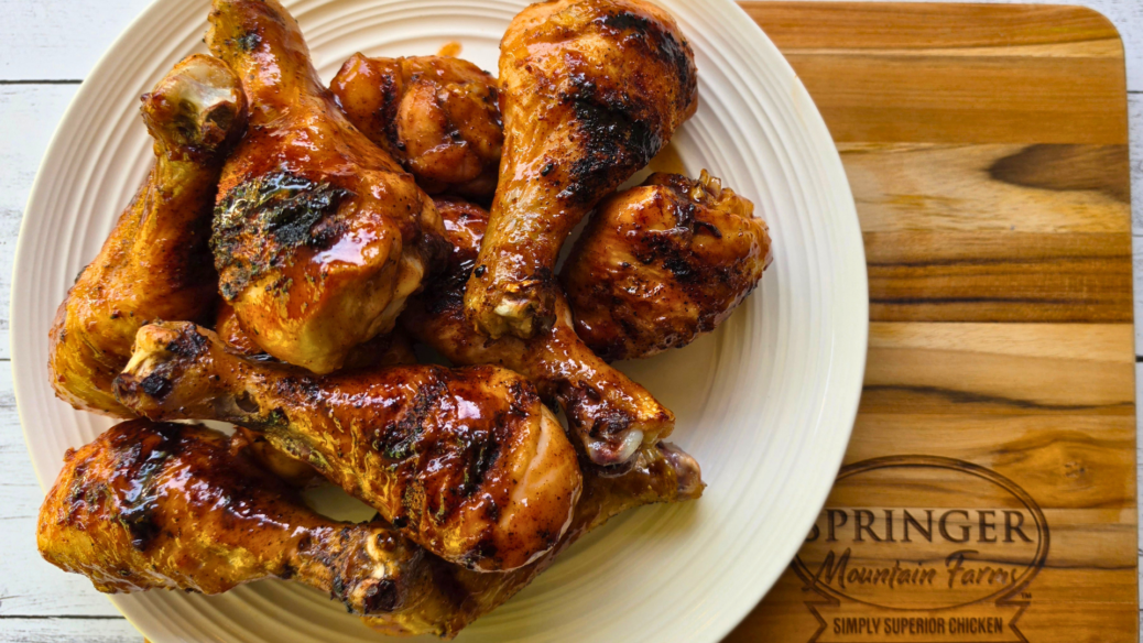 Smoky BBQ grilled chicken drumsticks piled on a plate, coated in a glossy barbecue sauce with visible char from the grill, served on a wooden board branded with Springer Mountain Farms