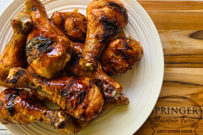 Smoky BBQ grilled chicken drumsticks piled on a plate, coated in a glossy barbecue sauce with visible char from the grill, served on a wooden board branded with Springer Mountain Farms
